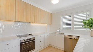 A modern kitchen with white cabinetry, a built-in oven, a dishwasher, and a window with blinds. - Beach Pines 5 - Fully Renovated North Kirra Beachfront - Min. 3 Night Stays (Bilinga)
