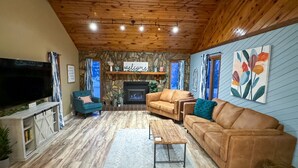 A cozy living room with a stone fireplace, wooden ceiling, and a large TV. - Theater Room | Hot Tub | Arcades | Great Location! (Spearfish)