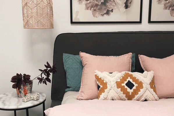 A bed with multiple pillows, a small round table with a plant, and a wall-mounted light fixture. - Joy Apartment 2 (Lorne)