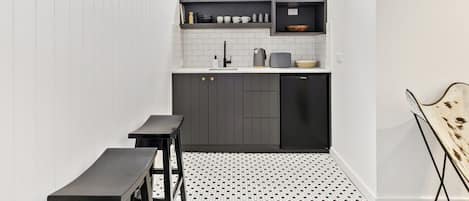 A compact kitchen with a built-in window, white tiled backsplash, dark cabinetry, and a small dining area with a bench.