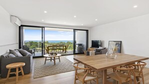 En moderne stue med et træspisebord, en grå sofa og en stor skydedør, der fører til en balkon med havudsigt.