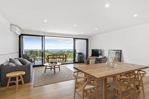 Ein modernes Wohnzimmer mit einem Holzesstisch, einer grauen Couch und einer großen Schiebeglastür, die auf einen Balkon mit Meerblick führt.