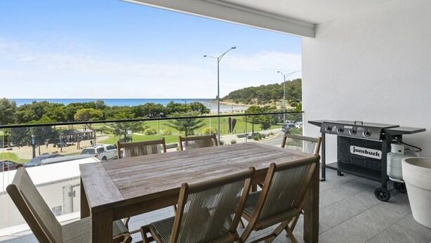 En balkon med et træbord og stole, en grill og udsigt til havet og grønne områder.