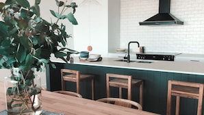 Una cocina moderna con encimera blanca, grifo negro y salpicadero verde. Hay una mesa de comedor de madera con sillas y un florero con hojas verdes.