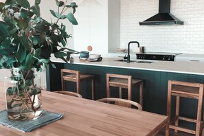 A modern kitchen with a white countertop, black faucet, and a green backsplash. There is a wooden dining table with chairs and a vase of green leaves.