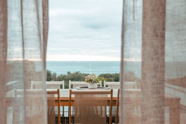 A dining area with a table and chairs, a potted plant, and a view of the sea and trees.