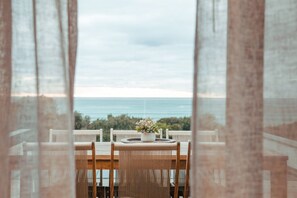 Ein Essbereich mit Tisch und Stühlen, einer Topfpflanze und Blick auf Meer und Bäume.