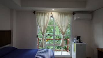 A bedroom with a bed, a window with curtains, an air conditioner, and a small table with a coffee maker.