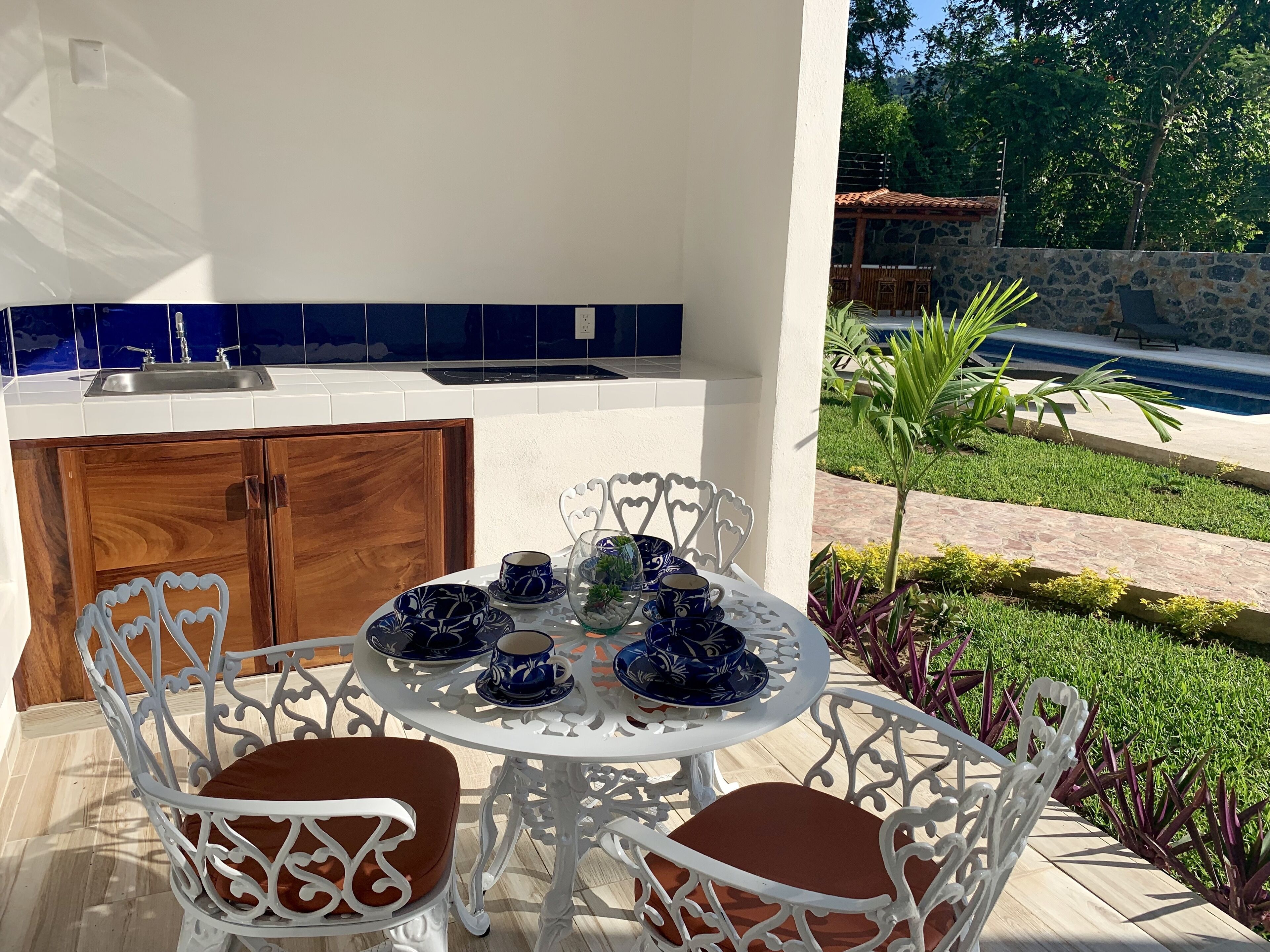 Une terrasse avec une table ronde, dressée avec de la vaisselle bleue et blanche, des chaises blanches, et une vue sur une piscine et un jardin.