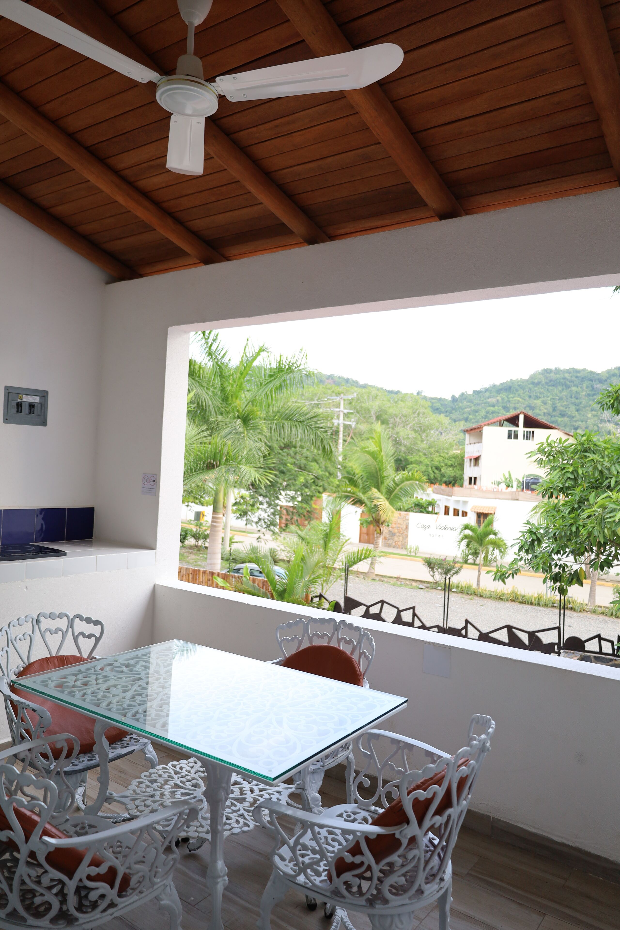 A room with a glass table and white chairs, a ceiling fan, and a view of a building and greenery outside.