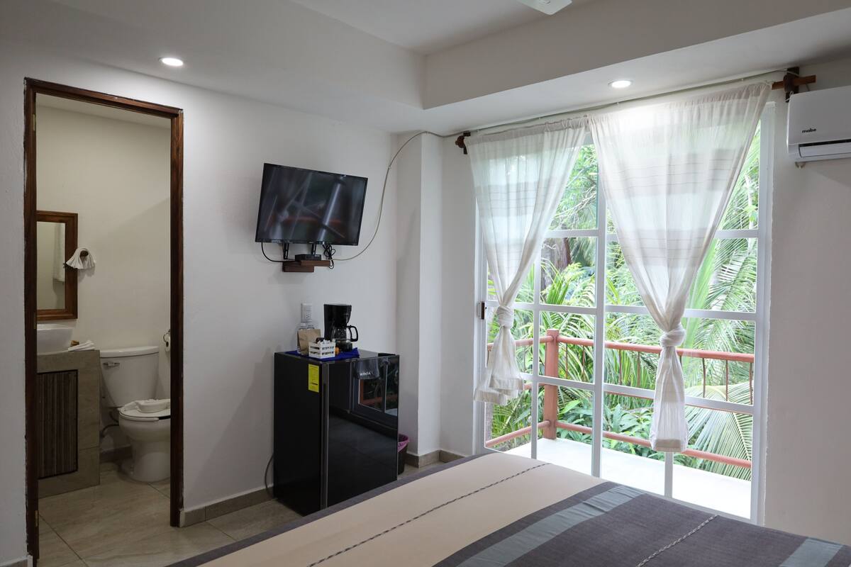Habitación de hotel con cama, televisor montado en la pared, kitchenette pequeña, ventana con cortinas y vista a zonas verdes.