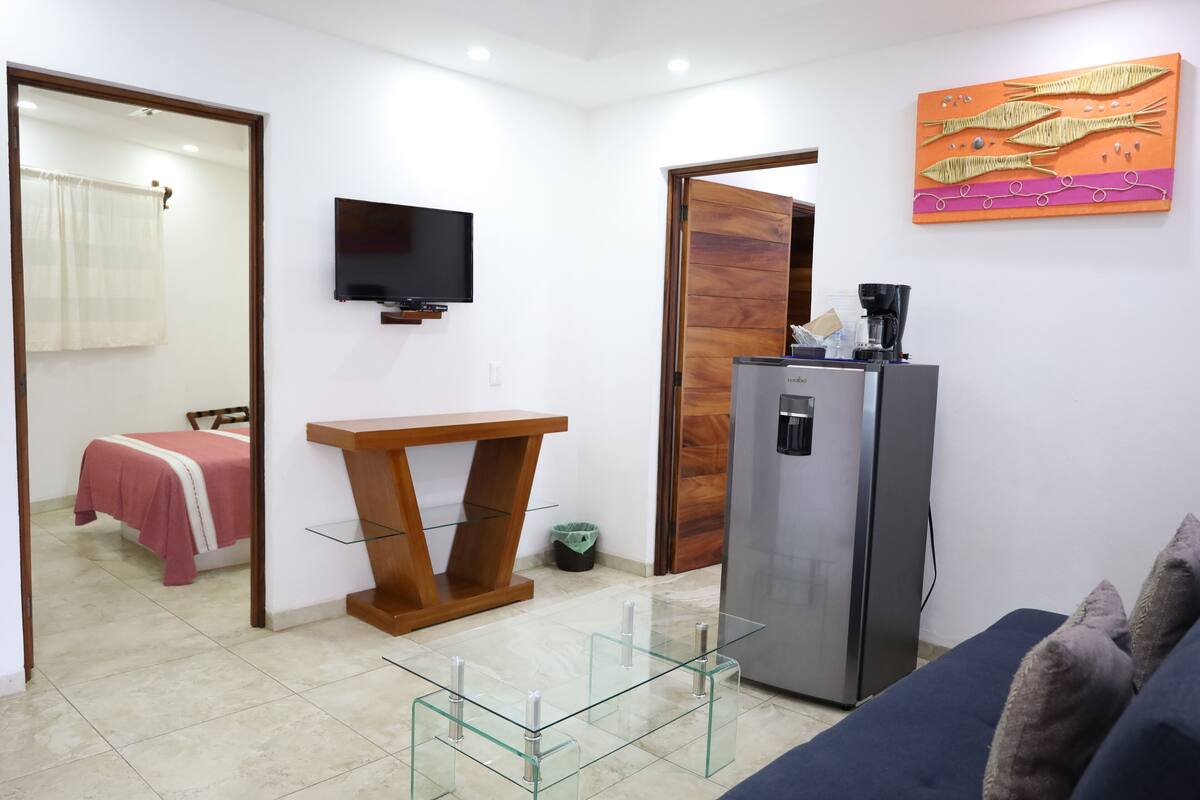 Un espacio habitable pequeño con cama, televisor, refrigerador, cafetera y una mesa de vidrio.