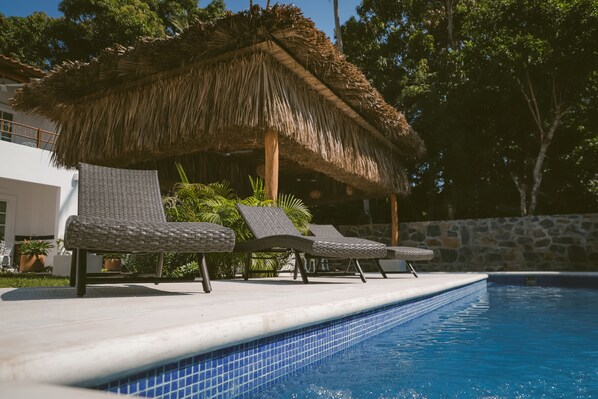 Zona junto a la piscina con sillones de descanso y una sombrilla de paja.