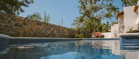 A clear swimming pool with stone walls and a tiled edge, surrounded by greenery and a building with a red roof.