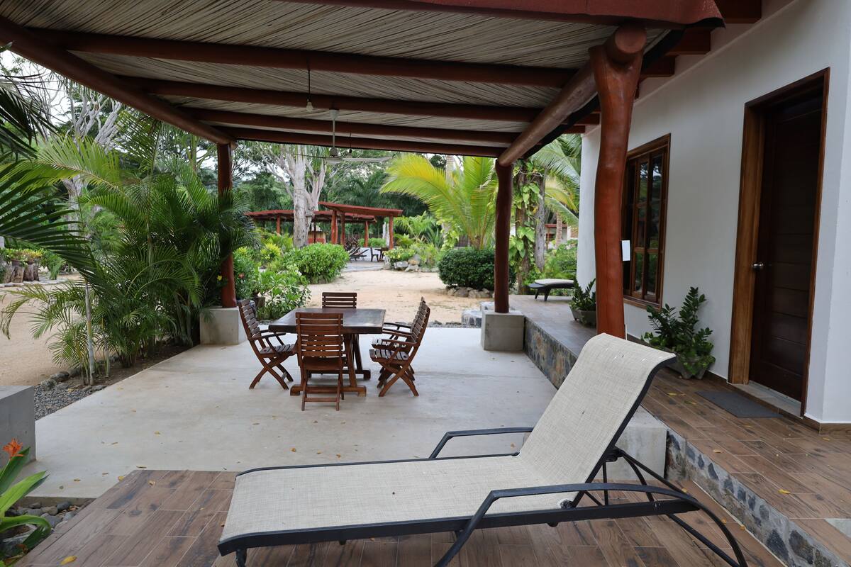 Un patio exterior con una mesa y sillas, rodeado de plantas y una pérgola de madera.