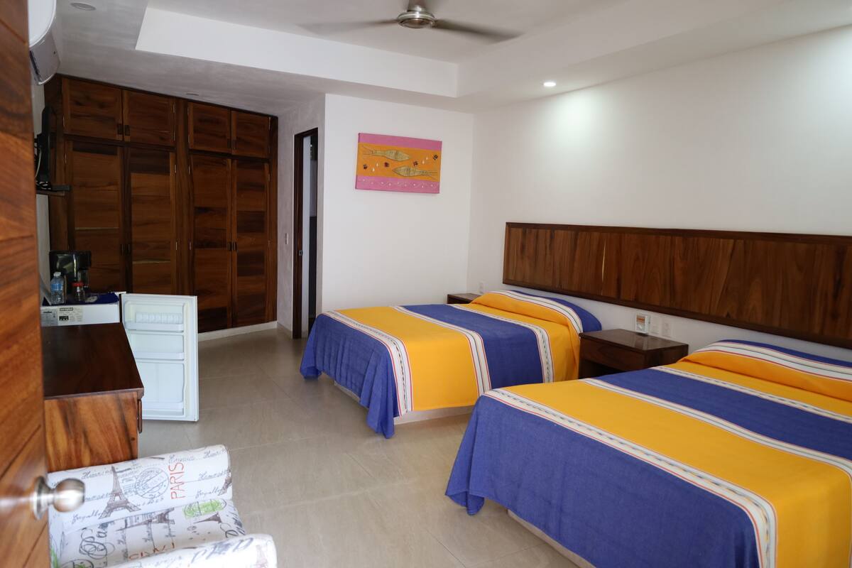Habitación de hotel con dos camas, un armario de madera, escritorio y una pequeña kitchenette.