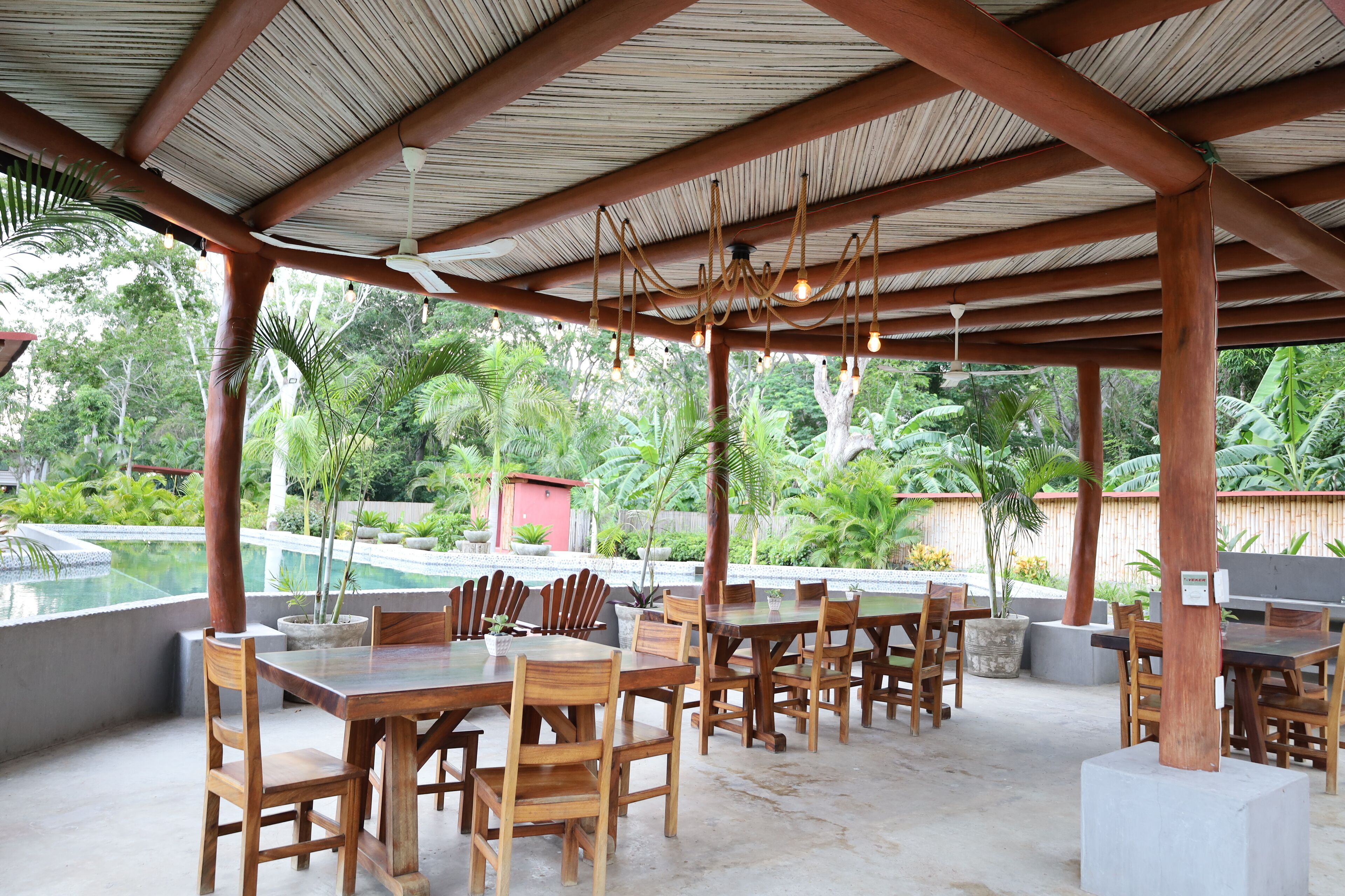 Une terrasse extérieure couverte, avec des tables et des chaises en bois, une piscine en arrière-plan et une végétation luxuriante.