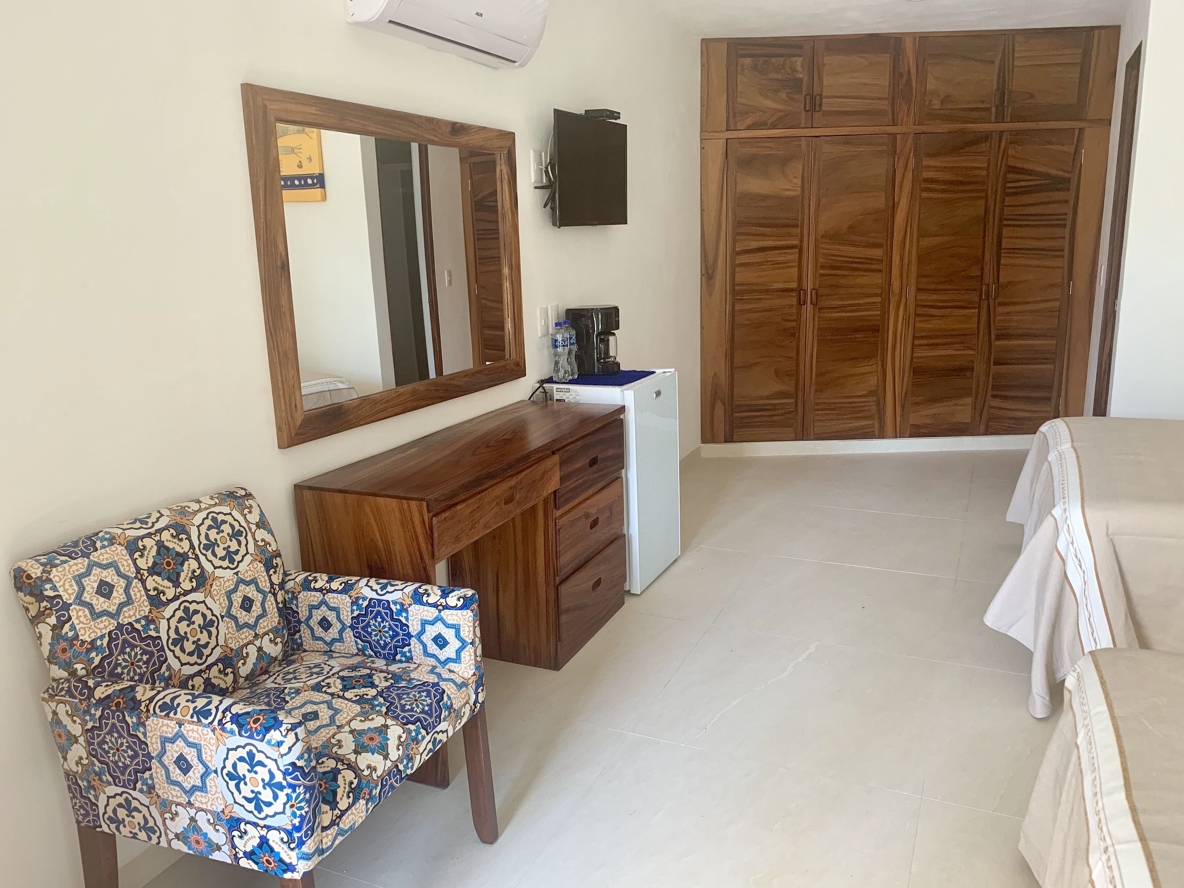 Une chambre à coucher avec une armoire en bois, un bureau avec un miroir, une chaise à motifs et un lit recouvert d’une couverture blanche.