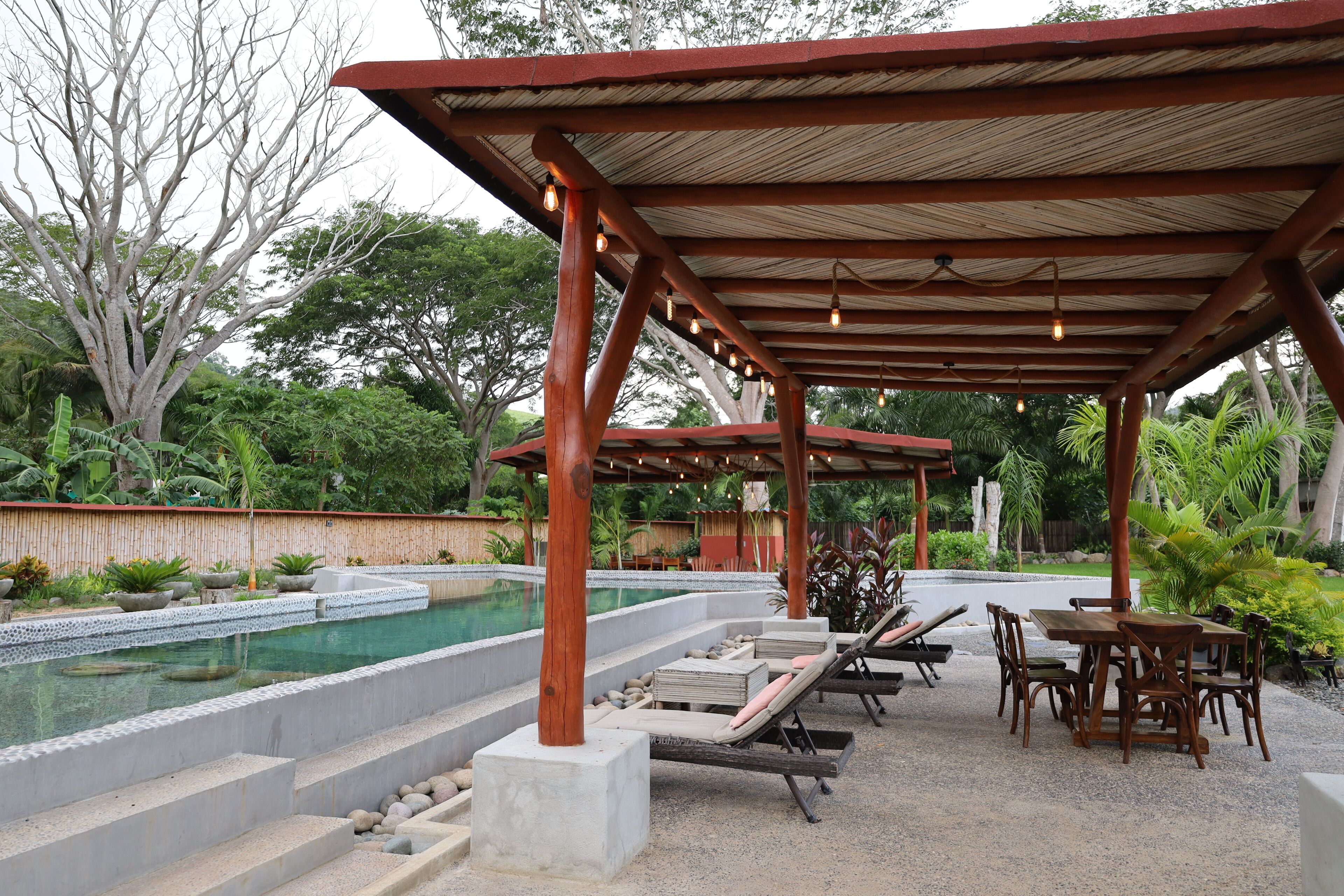 A covered outdoor area with a pool, lounge chairs, and a table with chairs.