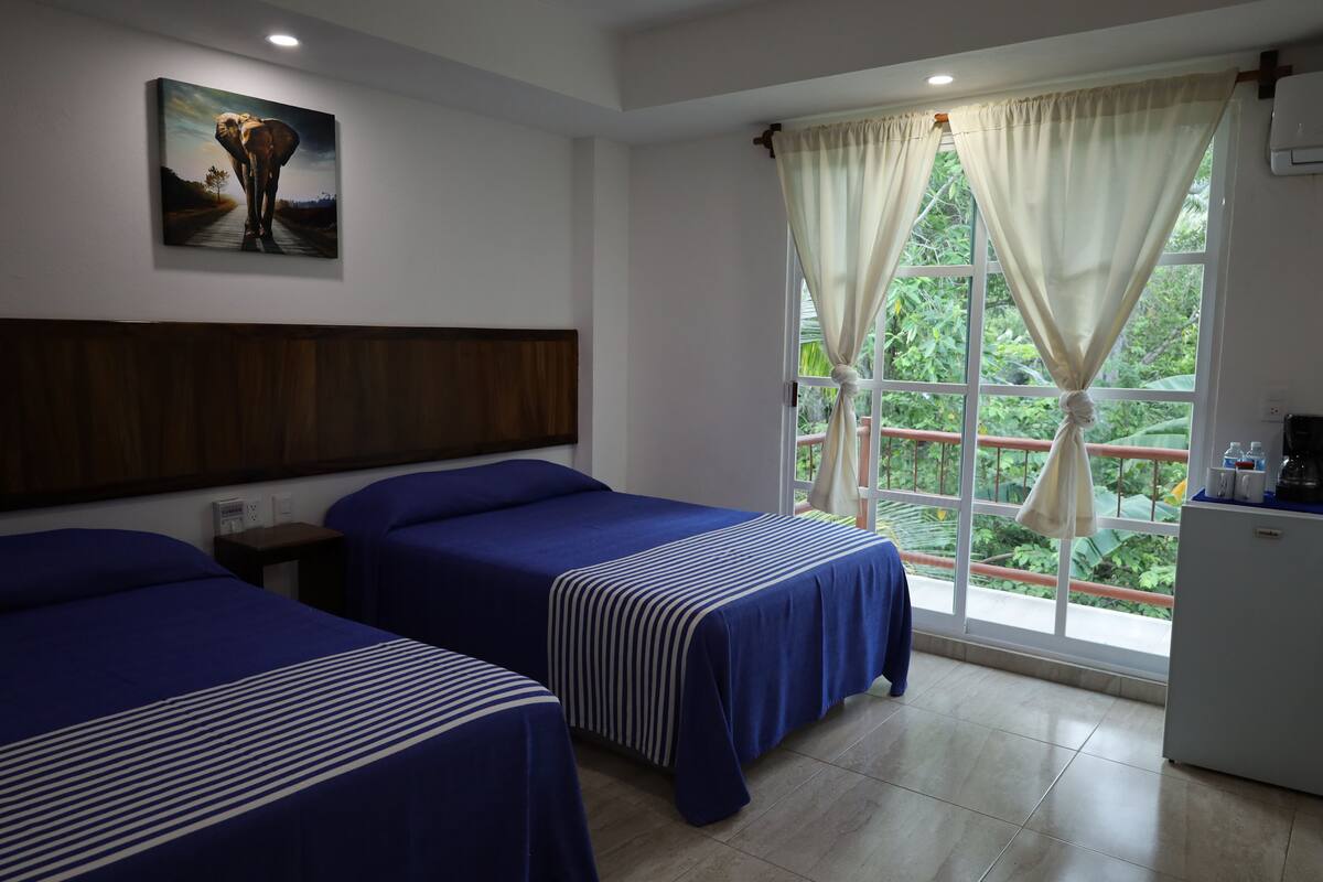 Una habitación de hotel con dos camas, un cuadro en la pared, una ventana con cortinas y un refrigerador pequeño.