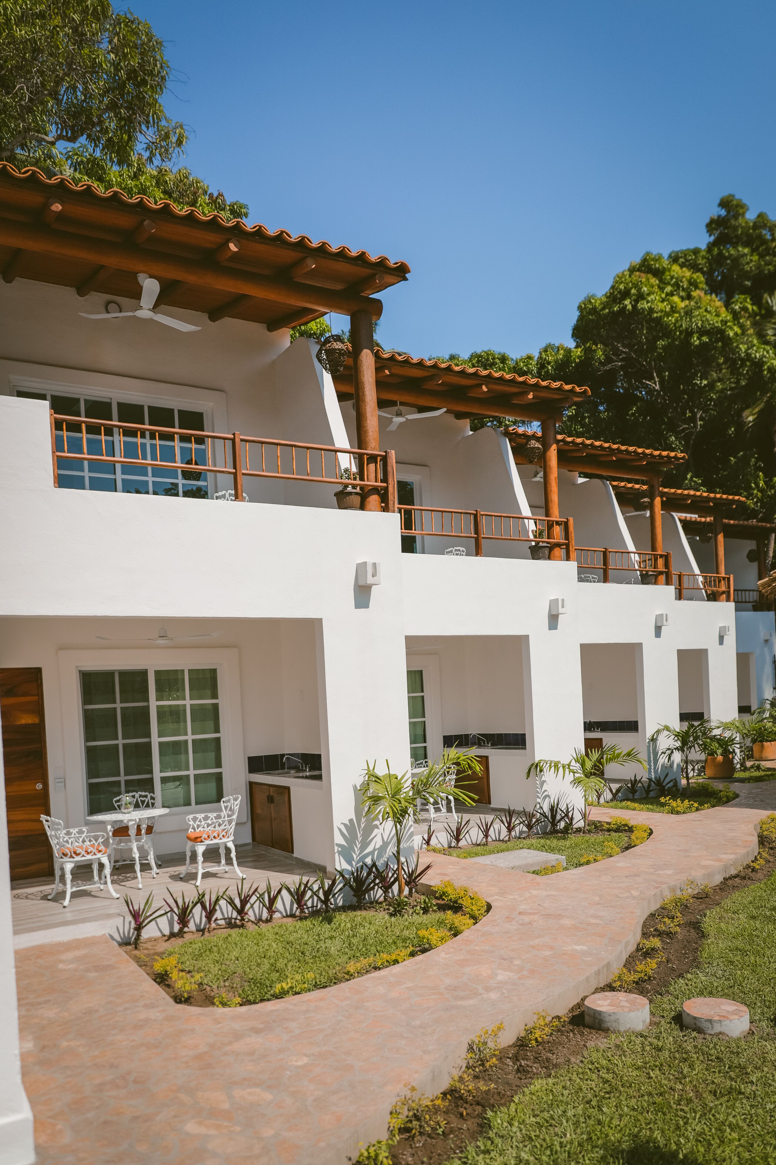 Un hôtel moderne doté d’un espace extérieur couvert avec des sièges, d’un sentier aménagé dans un jardin et d’une structure à deux étages avec des balcons.