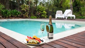 A poolside setting with a wooden deck, a plate of fruit, two glasses, and a bottle of champagne. - Luxury 4 bedroom, 4 bathroom apartment in Central Airlie with the best views (Airlie Beach)