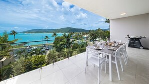 A balcony with a dining table set for four, overlooking a coastal view with a clear sky and distant mountains. - Luxury 4 bedroom, 4 bathroom apartment in Central Airlie with the best views (Airlie Beach)
