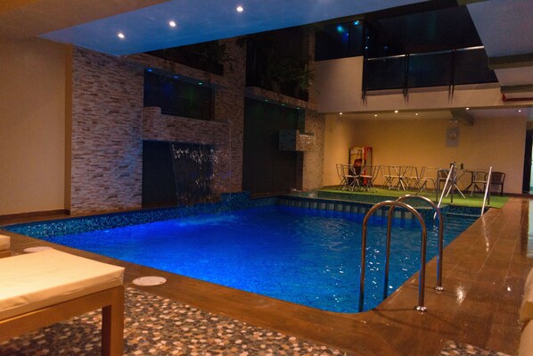 An indoor swimming pool with a brick wall, a waterfall feature, and a seating area with a table. - Hotel Paradis Suite - Cusco (Cusco)