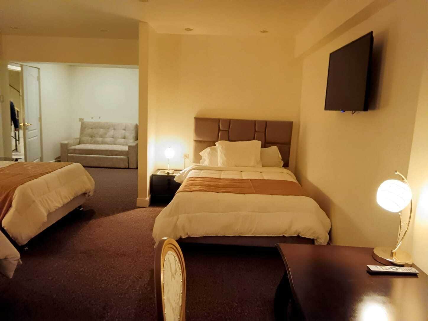 Quarto de hotel com duas camas, televisão na parede, mesa de cabeceira com abajur e uma cadeira no canto.
