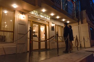 Entrada de un hotel con una barrera de cadena, un letrero que dice 'HOTEL PARADIS' y el interior visible a través de las puertas de vidrio.