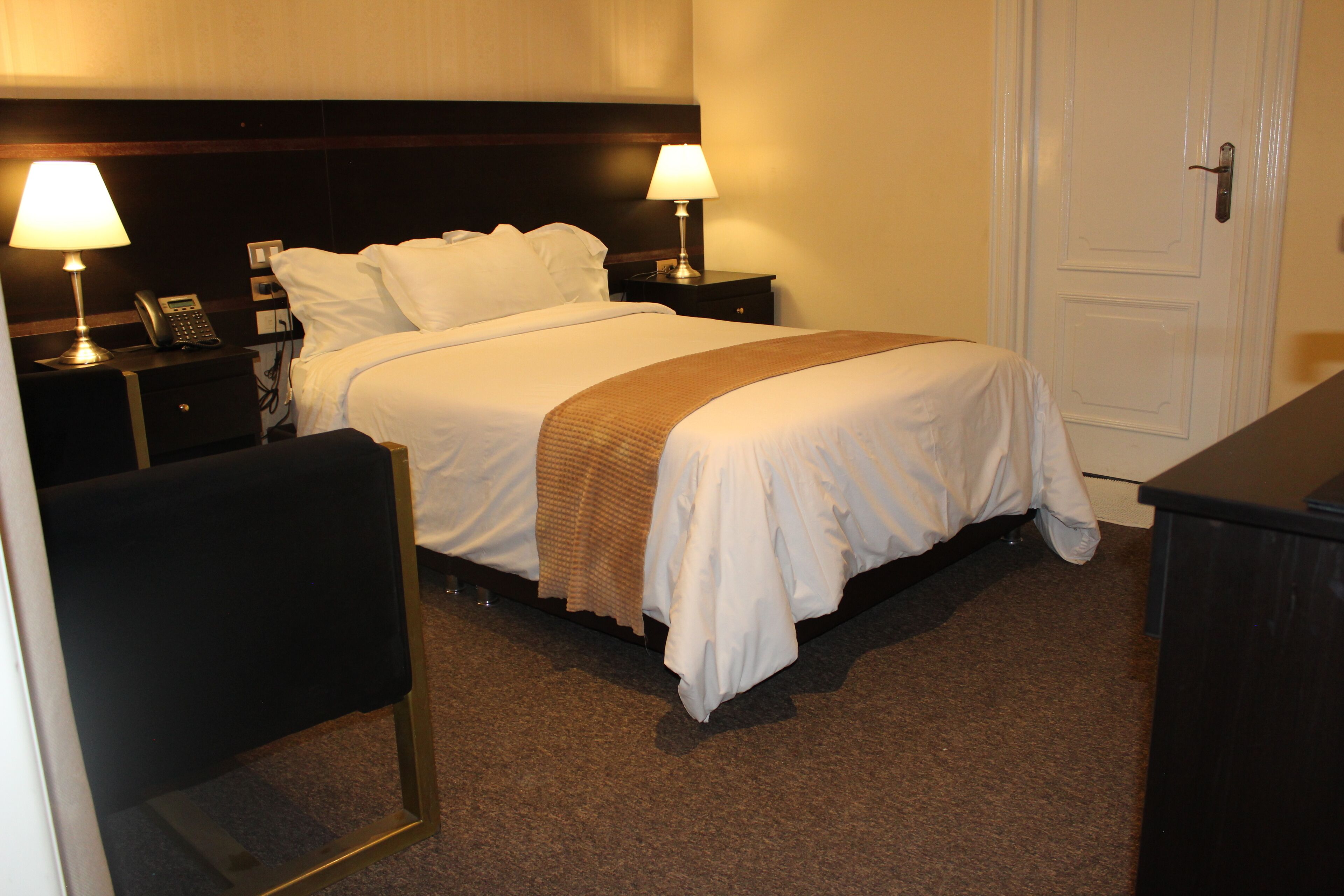Quarto de hotel com uma cama, duas mesas de cabeceira com abajures, um telefone e uma porta.