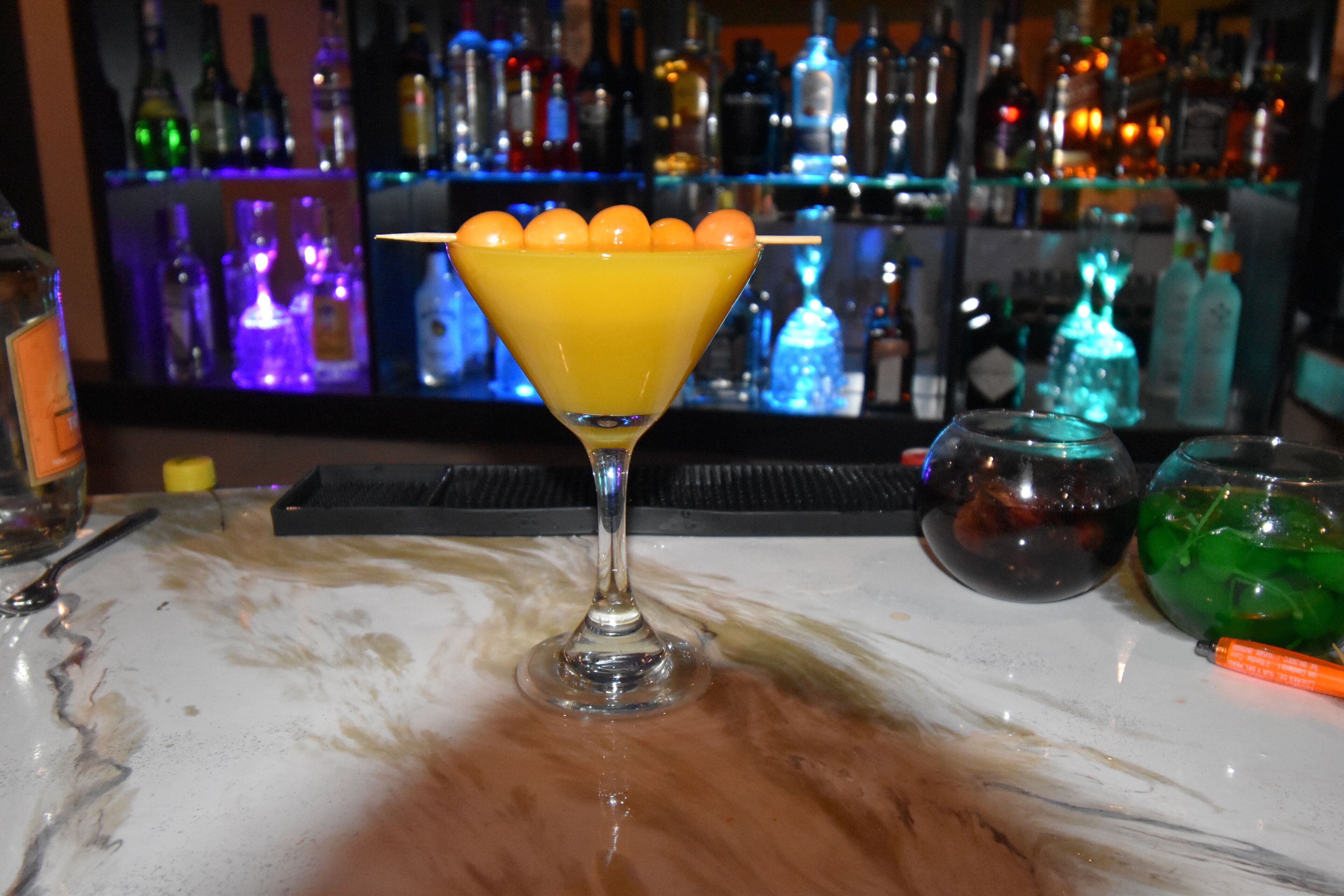 A bar setting with a martini glass with a lemon garnish, a green cocktail, and a bar area with various bottles.