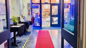 A hotel reception area with a red carpet leading to the entrance, a reception desk, and a view of the street outside.