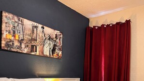 A hotel room with a bed, pillows, a blanket, a remote control, a painting on the wall, and a red curtain.