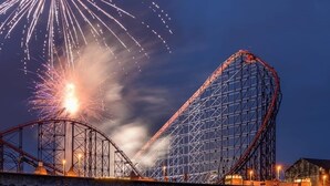 A roller coaster with a steep drop, surrounded by a body of water reflecting lights, and fireworks in the sky.