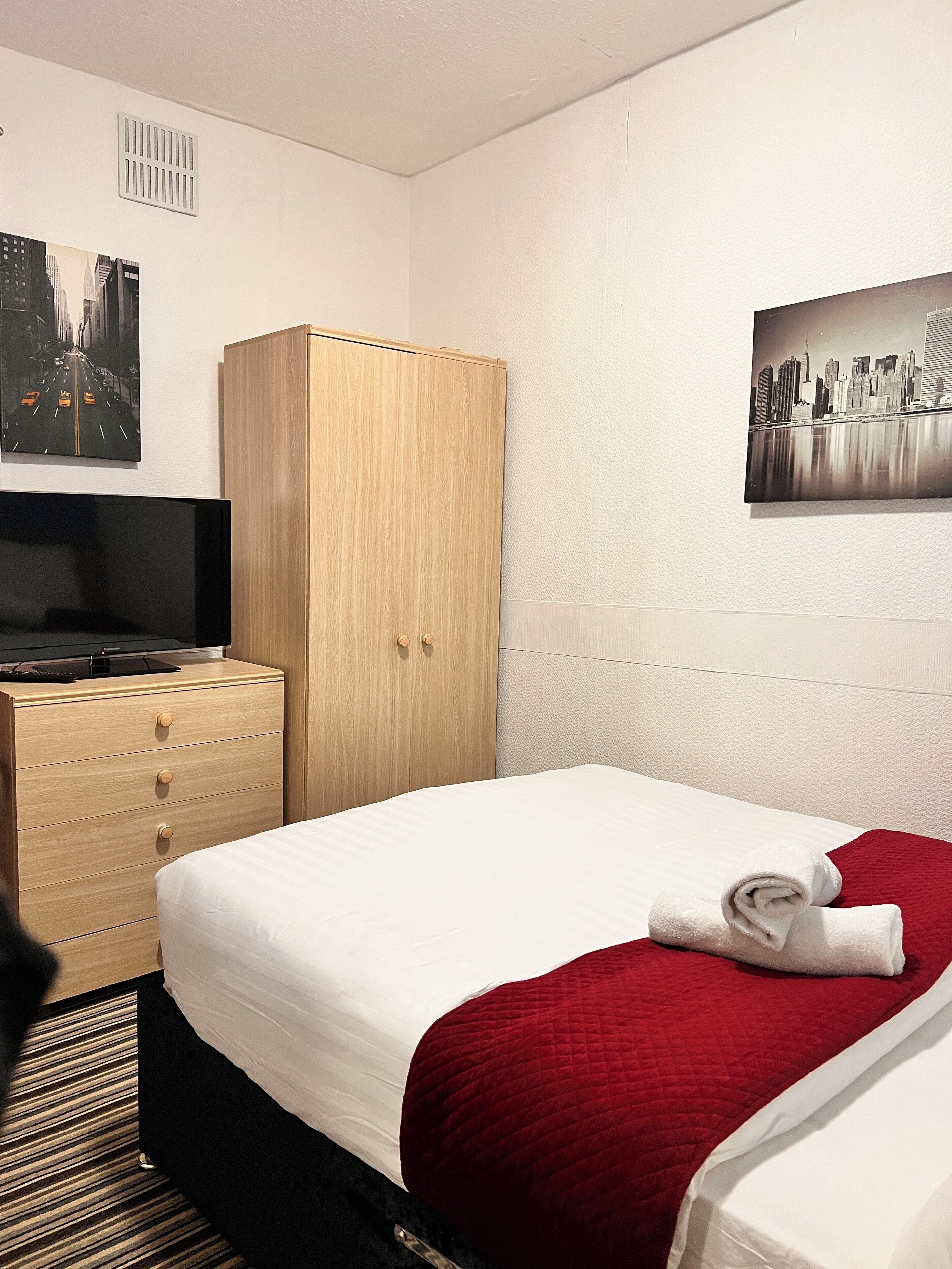 A hotel room with a bed, a wooden wardrobe, a television, and two cityscape paintings on the walls.