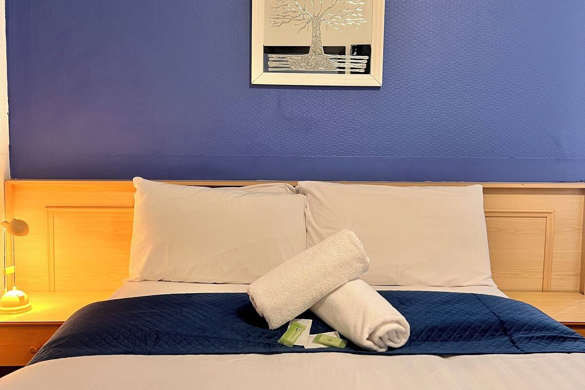 A neatly made bed with white and blue bedding, a rolled white towel, and a framed artwork on the wall.
