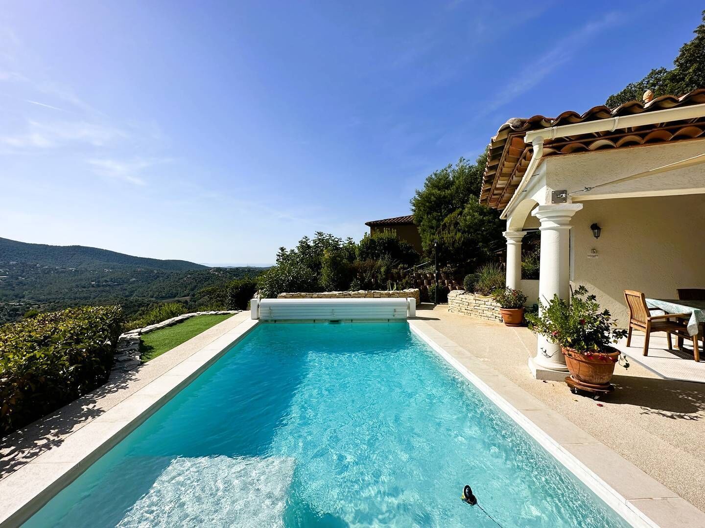 A rectangular swimming pool with clear blue water, surrounded by a stone deck and a white wall with columns. There is a view of a mountainous landscape in the background.