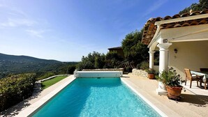 Una piscina rectangular con agua azul cristalina, rodeada por una terraza de piedra y un muro blanco con columnas. Al fondo, se ve un paisaje montañoso.
