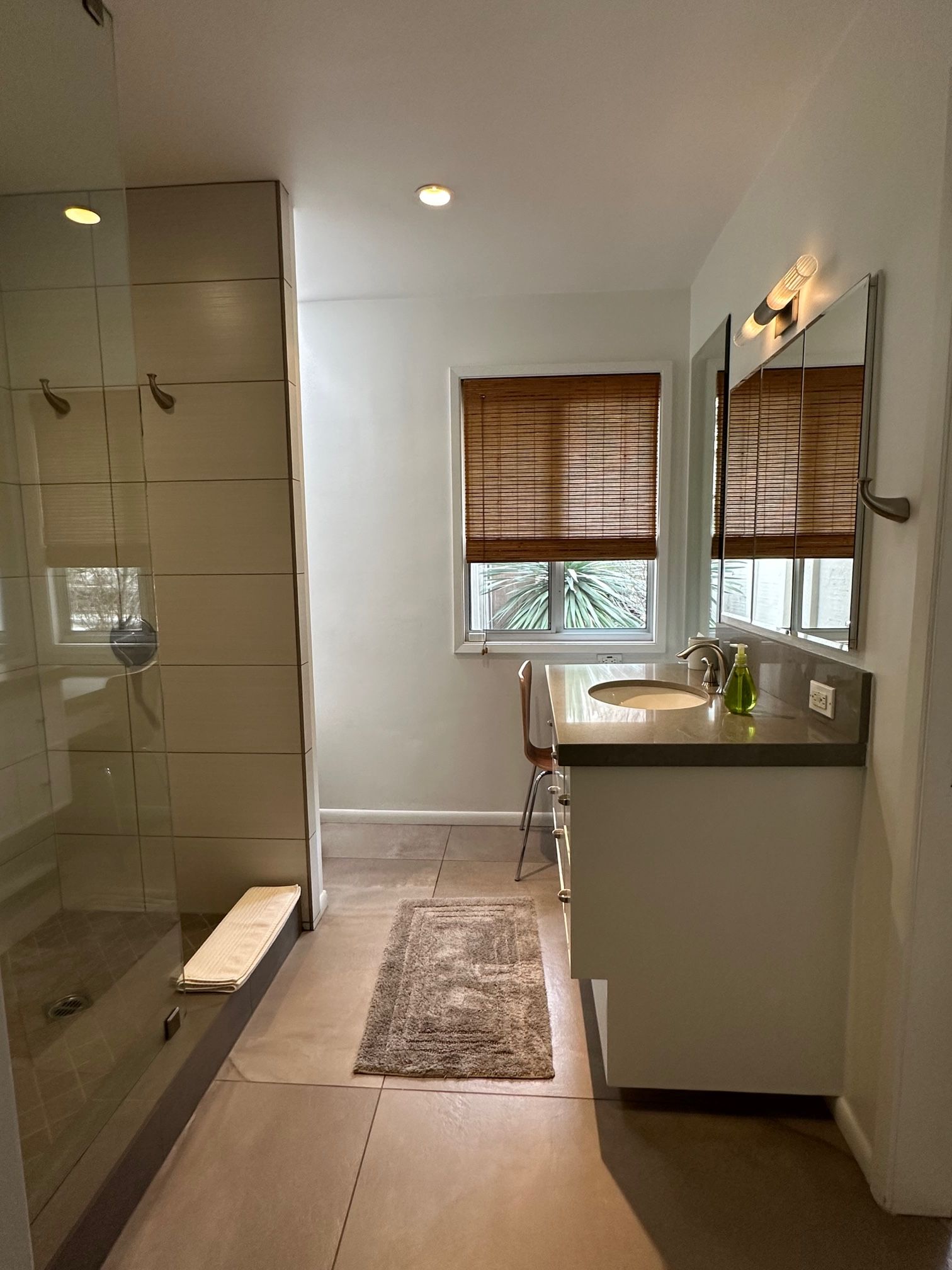 A bathroom with a glass shower enclosure, a sink with a mirror, and a window with blinds.