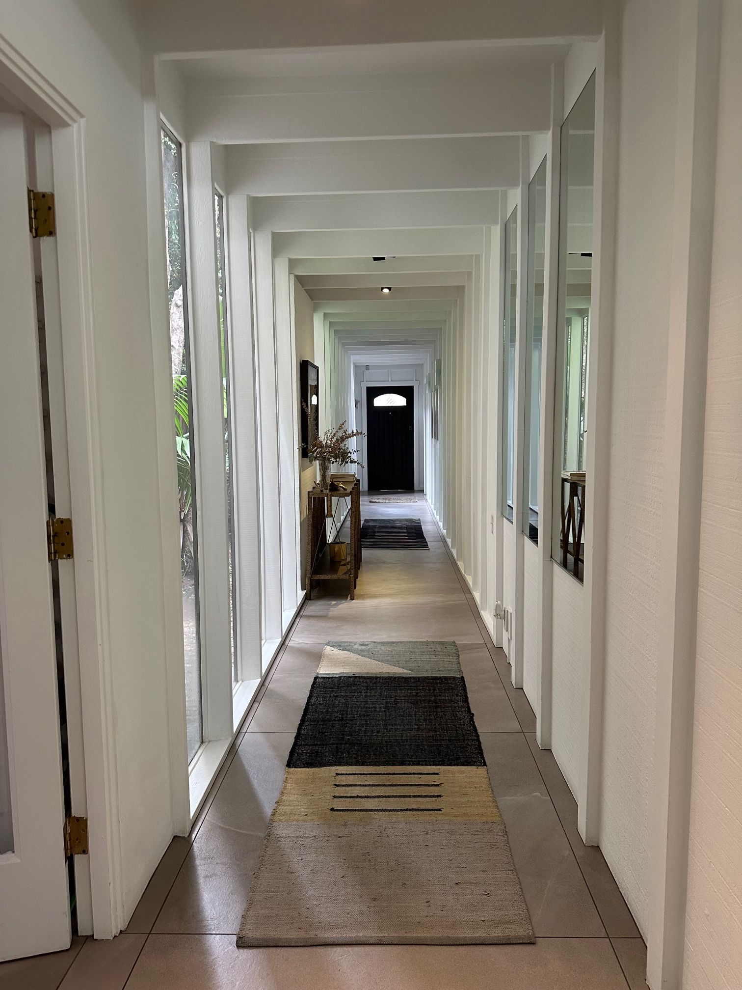 A long, narrow hallway with white walls and a tiled floor. There are large windows on both sides, allowing natural light to enter. A wooden bench with a vase of dried flowers is placed in the center of the hallway.