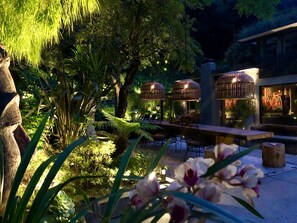 Un espace extérieur aménagé avec des tables et des chaises, entouré d’une végétation luxuriante et illuminé par un éclairage décoratif.