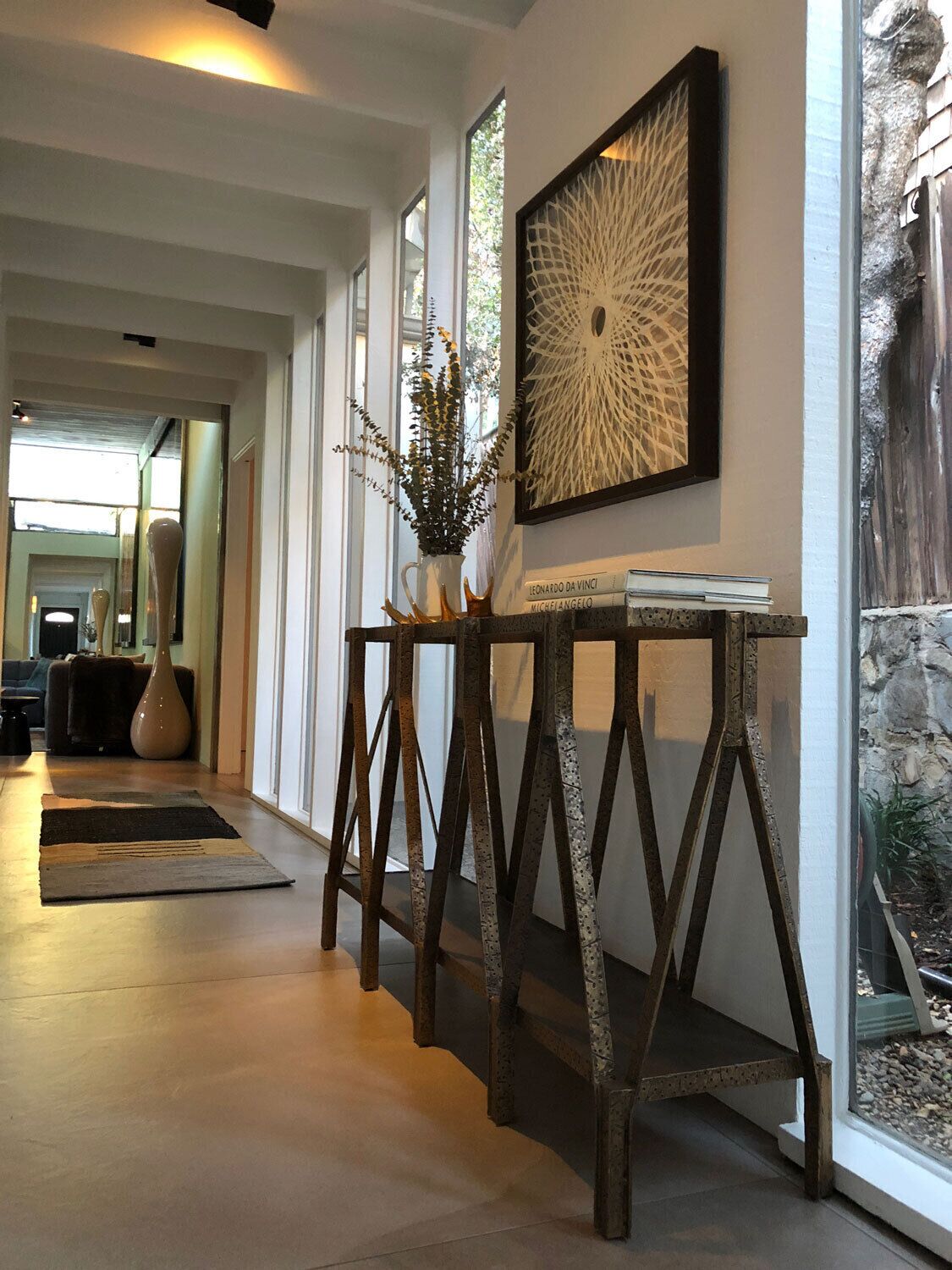 Un interior moderno con un largo pasillo, una mesa decorativa con un florero y una obra de arte enmarcada en la pared.