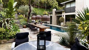 Un espace piscine extérieur moderne, doté d’une table et de chaises en verre, entouré d’une végétation luxuriante et bordé d’un bâtiment contemporain.