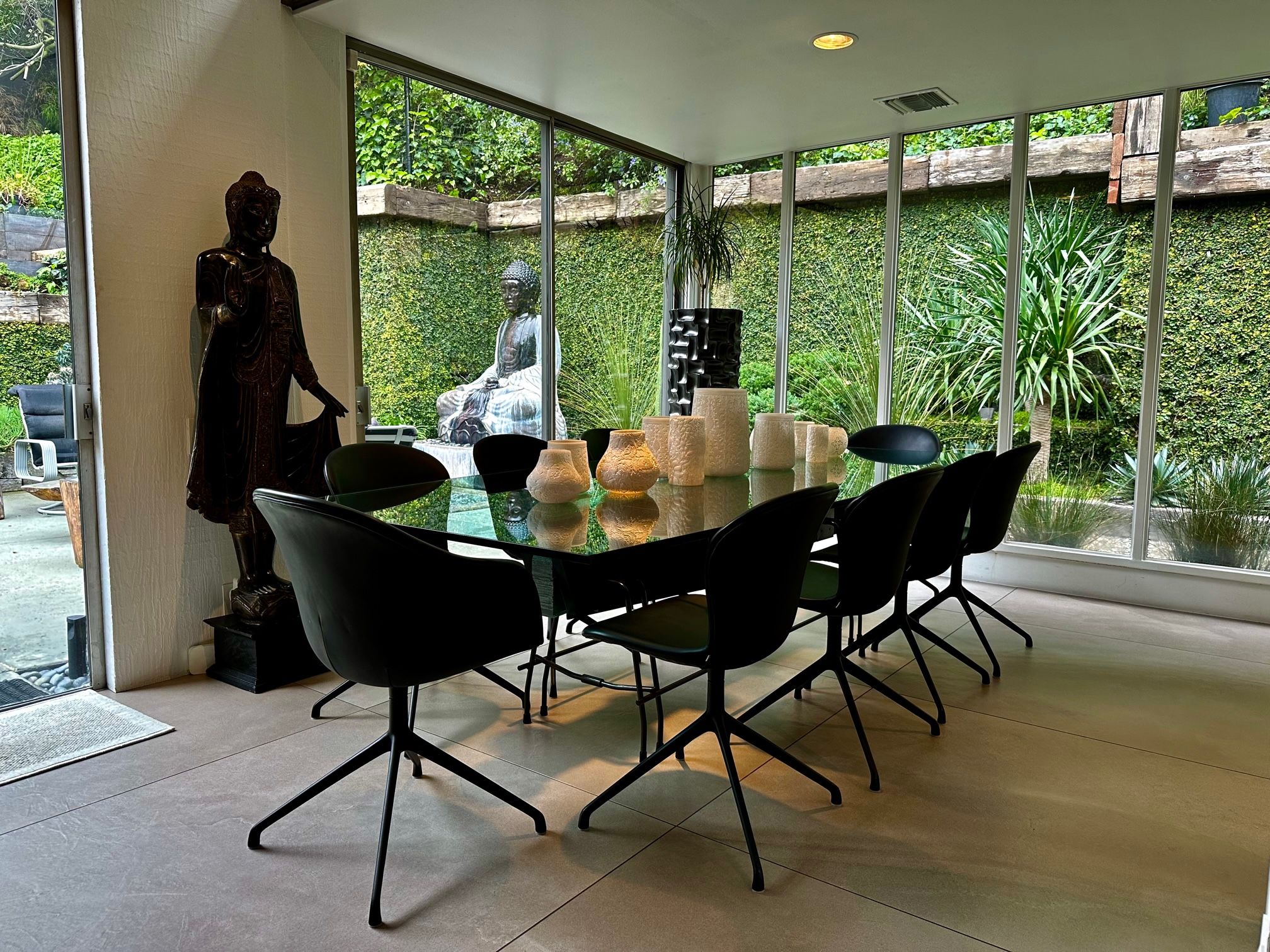 A modern dining area with a glass table, black chairs, and a view of a garden through large windows.
