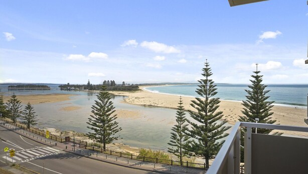 A coastal view with a beach, a road with pedestrian crossing, and a bridge in the distance.
