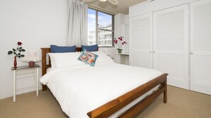 A bedroom with a wooden bed, white bedding, a nightstand with a vase of flowers, and a window with a view of buildings.