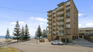 A multi-story residential building with balconies, located near a beach with trees and a clear sky.
