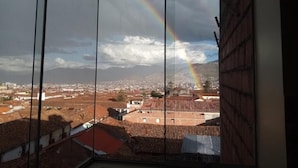 A view through a window with a rainbow, buildings, and mountains in the background. - Marvelous Plaza (Cusco)