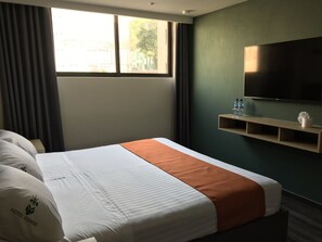 A hotel room with a bed, a TV mounted on the wall, a window with curtains, and a shelf with water bottles. - Hotel Conde Alameda - CDMX (Mexico City)